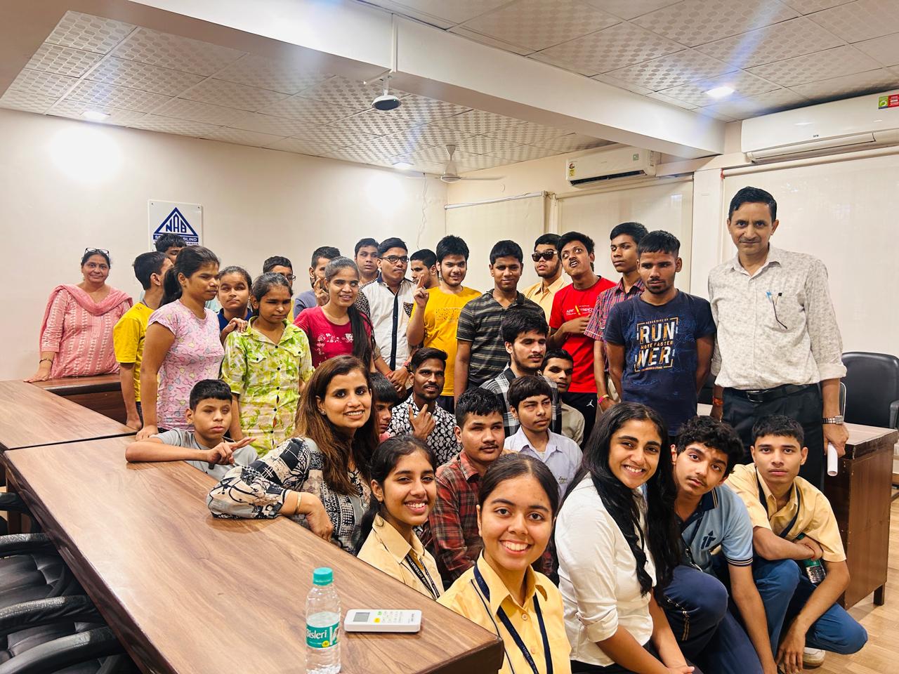The image shows a group of people gathered in a room, for a group photo. There are about 30 individuals, including both adults and children. Most of them are standing, while a few are sitting at a long wooden table in the foreground. The group is diverse, with both boys and girls, and they appear to be of school students. Some of the individuals are smiling at the camera. The room has a ceiling with recessed lighting and air conditioning units. On the wall in the background, there is a sign with the letters 'NAB' on it. A bottle of water and a remote control are visible on the table in the foreground. The atmosphere seems friendly and informal.